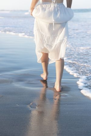 Bare feet walking along the shoreline in a white dress leaving footprints in wet sand.