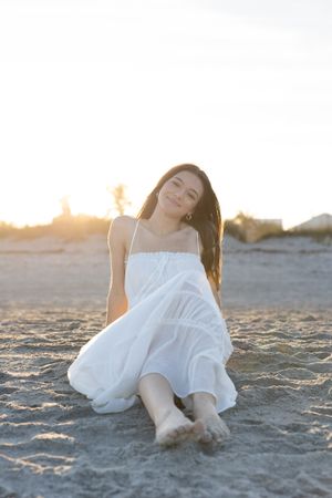 Woman in white dress relaxing on sandy beach at sunset with dreamy expression.