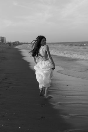 Figure in flowing white dress running along beach shoreline in black and white.