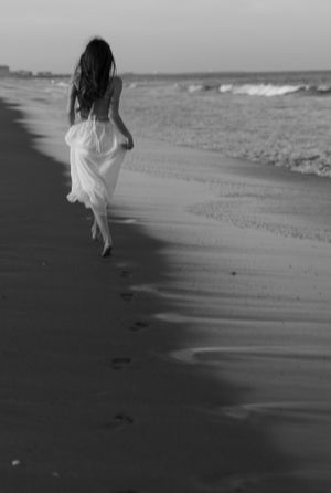 Person walking along beach leaving footprints in wet sand at sunset in black and white.