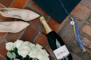 Graduation celebration items including white heels, champagne, roses and tassel on brick surface.