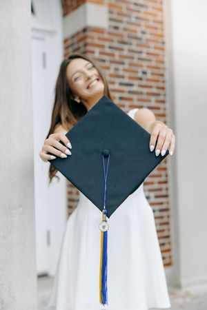 Graduate holding mortarboard cap with blue tassel against brick wall.