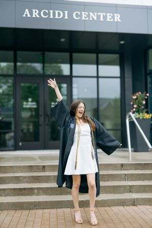 Graduate celebrating in cap and gown outside Arcidi Center building.