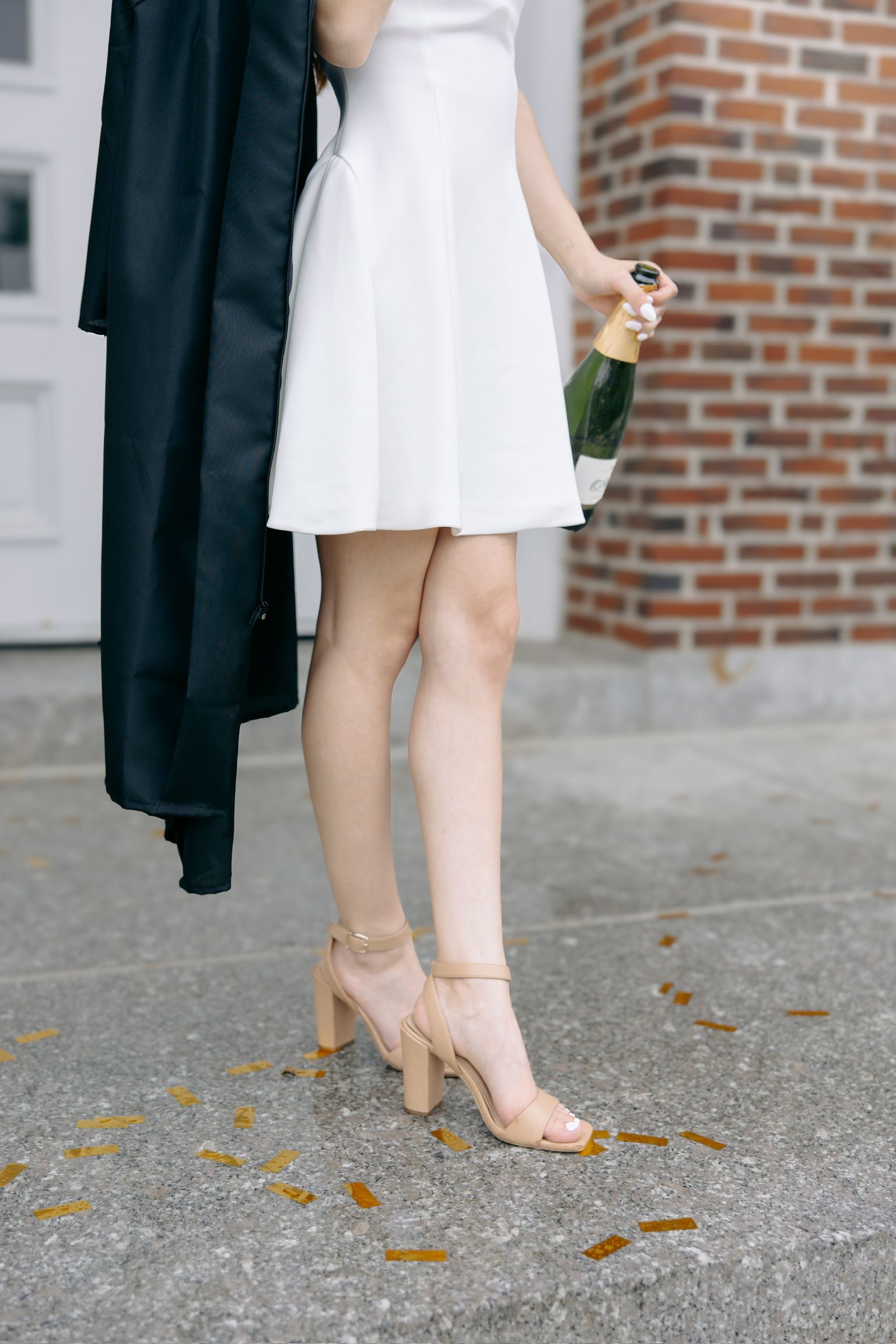 Close-up of graduate's white dress, graduation gown and nude heels on concrete.
