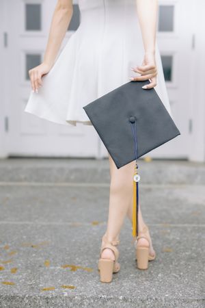 Person holding graduation cap and tassel while wearing heels on concrete ground.