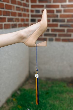 Graduation cap and tassel hanging from a hand against a brick wall background.