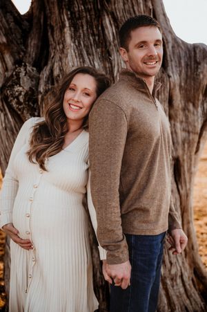 Smiling pregnant couple posing together in front of a large tree, woman in white dress holding baby bump.