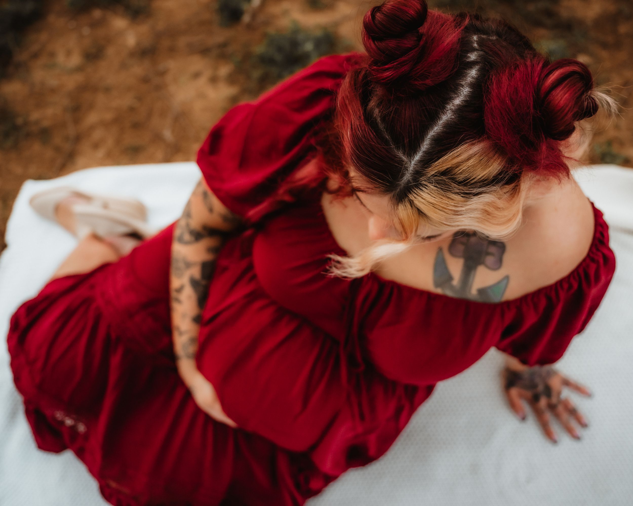 Woman in red dress with tattoos sitting gracefully, red hair in buns, viewed from above on white surface.