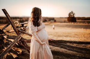 A person in a flowing white dress cradles their pregnant belly while standing near a wooden fence at sunset.