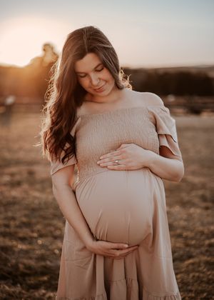 A person in a beige sweater cradles their pregnant belly during a golden hour maternity photoshoot in a field.