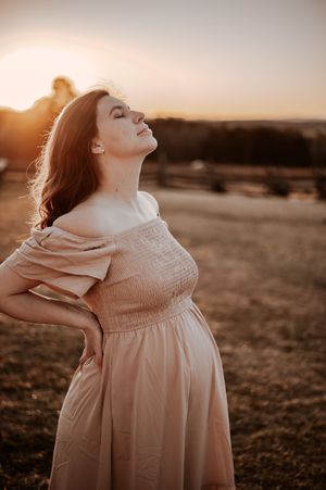 A pregnant woman in a flowing pink dress stands in golden sunlight at sunset, tilting her head back with closed eyes.