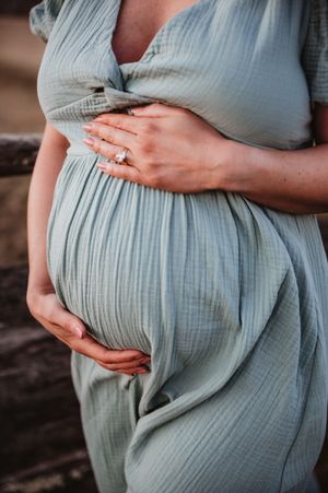 Pregnant woman in sage green dress cradling baby bump, wearing engagement ring, maternity photography outdoors.