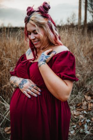 Smiling pregnant woman in red dress with tattoos and pink hair buns poses outdoors in a field.