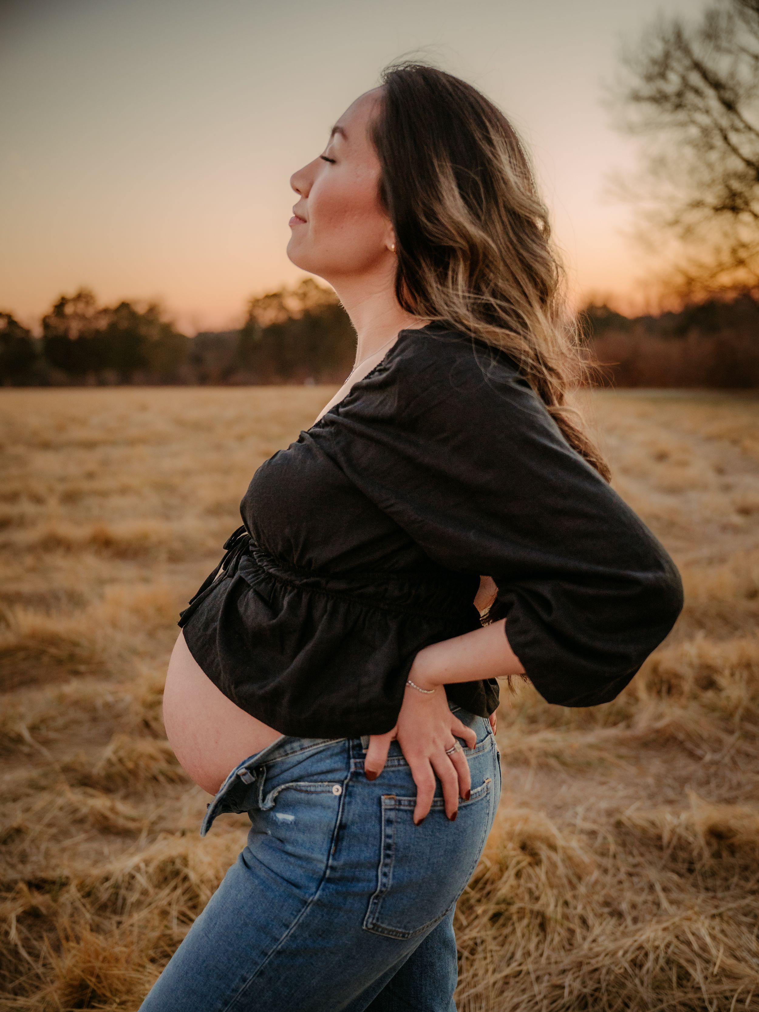 Maternity photos in golden field at sunset showing woman in black top and jeans in Stafford VA.