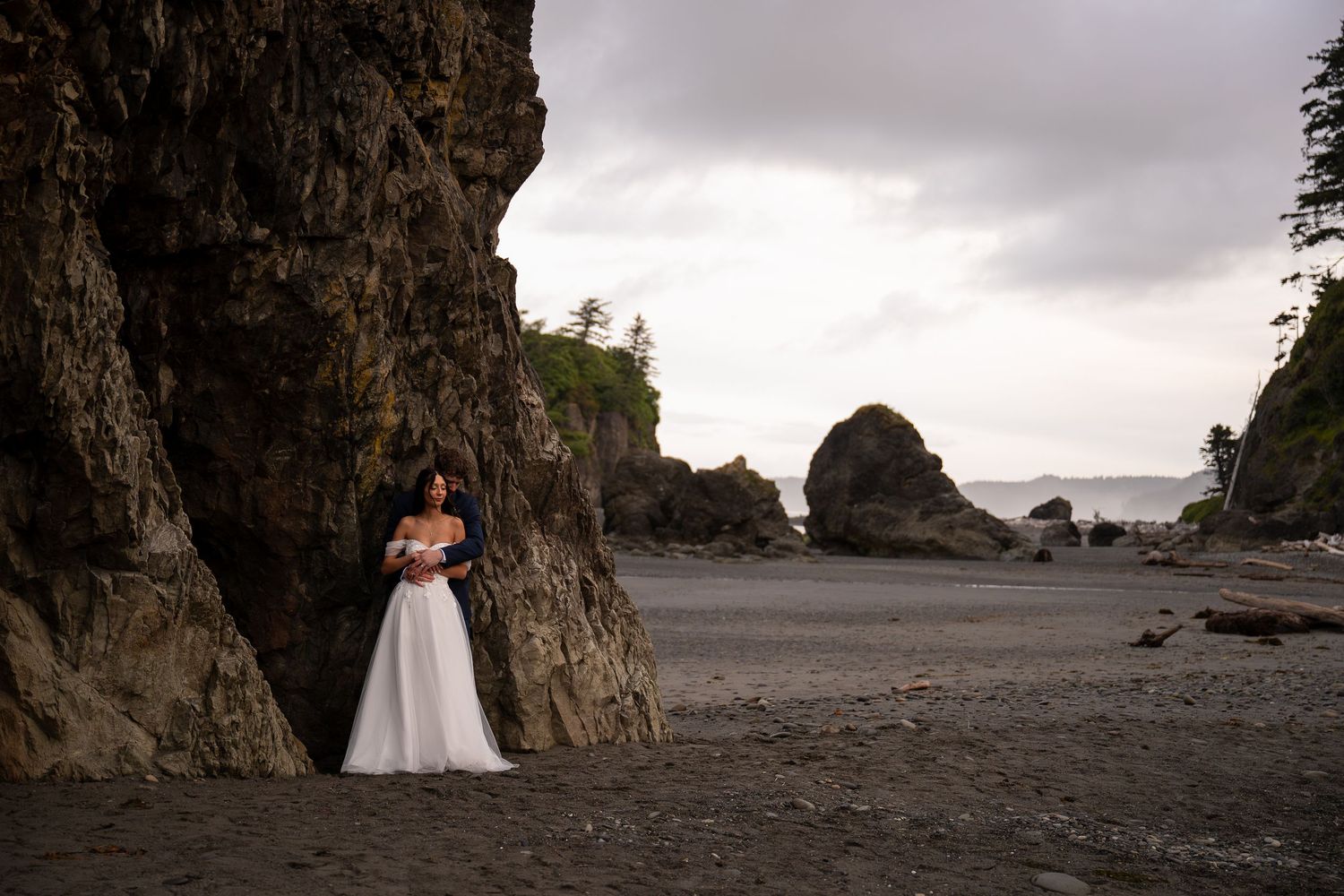 Newlyweds cuddle to stay warm in their wedding attire on a rugged Pacific Northwest beach.