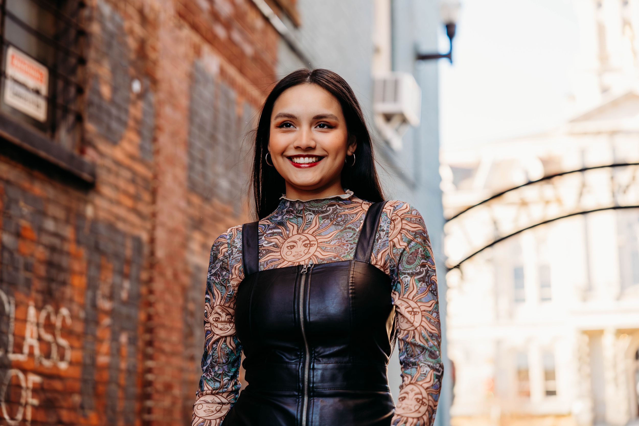 A person in a leather vest and lace sleeves poses against a brick wall in an urban setting.