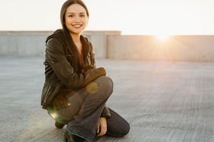 A person in casual wear sits and smiles on a concrete surface during golden hour lighting.