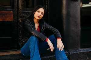 Person in black leather jacket and blue jeans sits casually on outdoor steps in moody lighting.