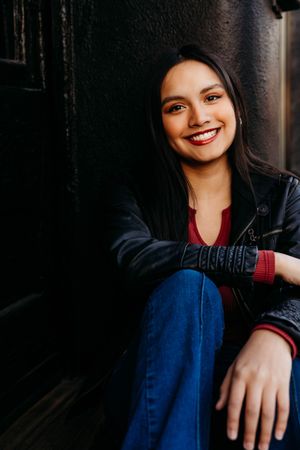 Portrait of someone in a black leather jacket and blue jeans sitting casually against a dark wall with a warm smile.