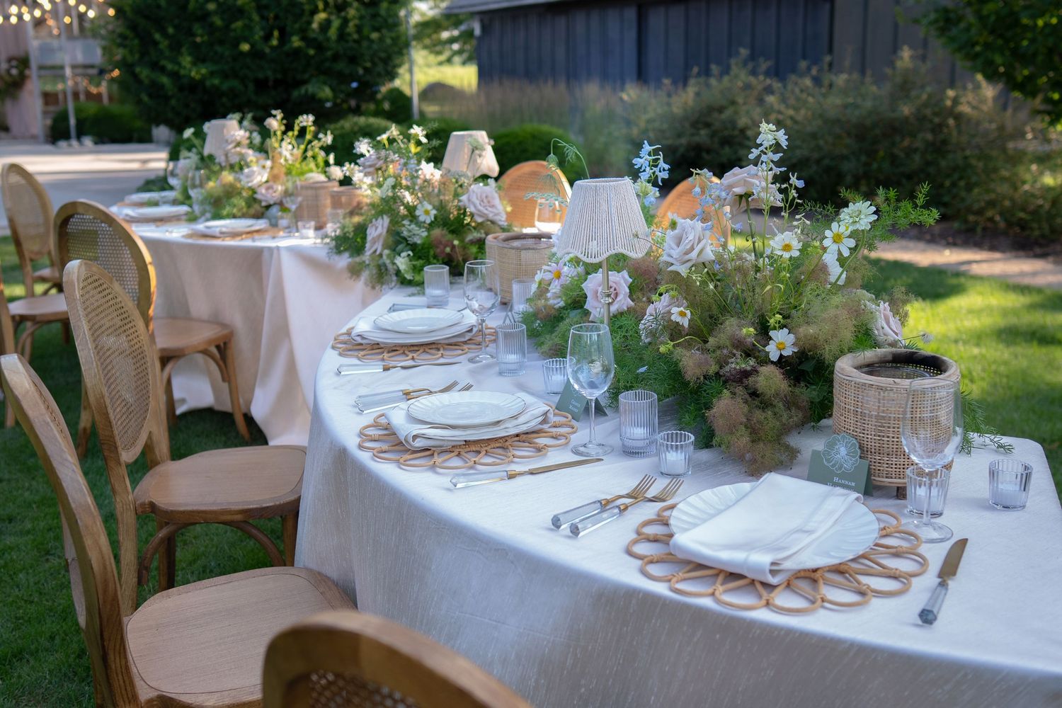 Elegant outdoor table setting with white linens, wooden chairs, and delicate floral centerpieces in a garden setting.