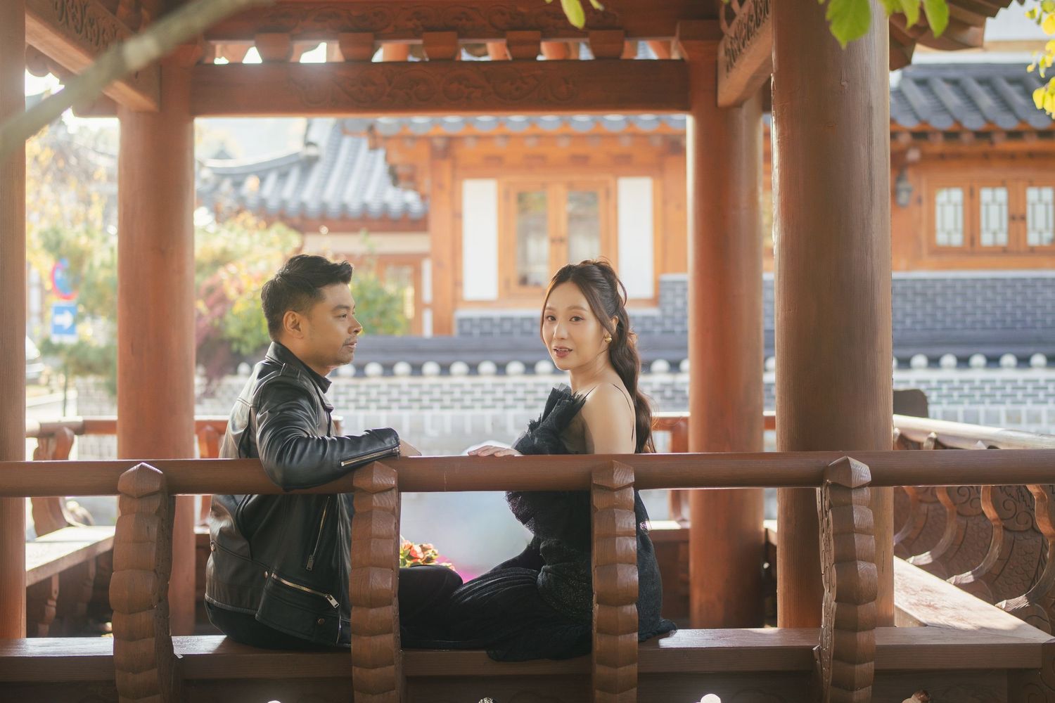 Two people sit and talk in a traditional Asian covered walkway with ornate wooden architecture.