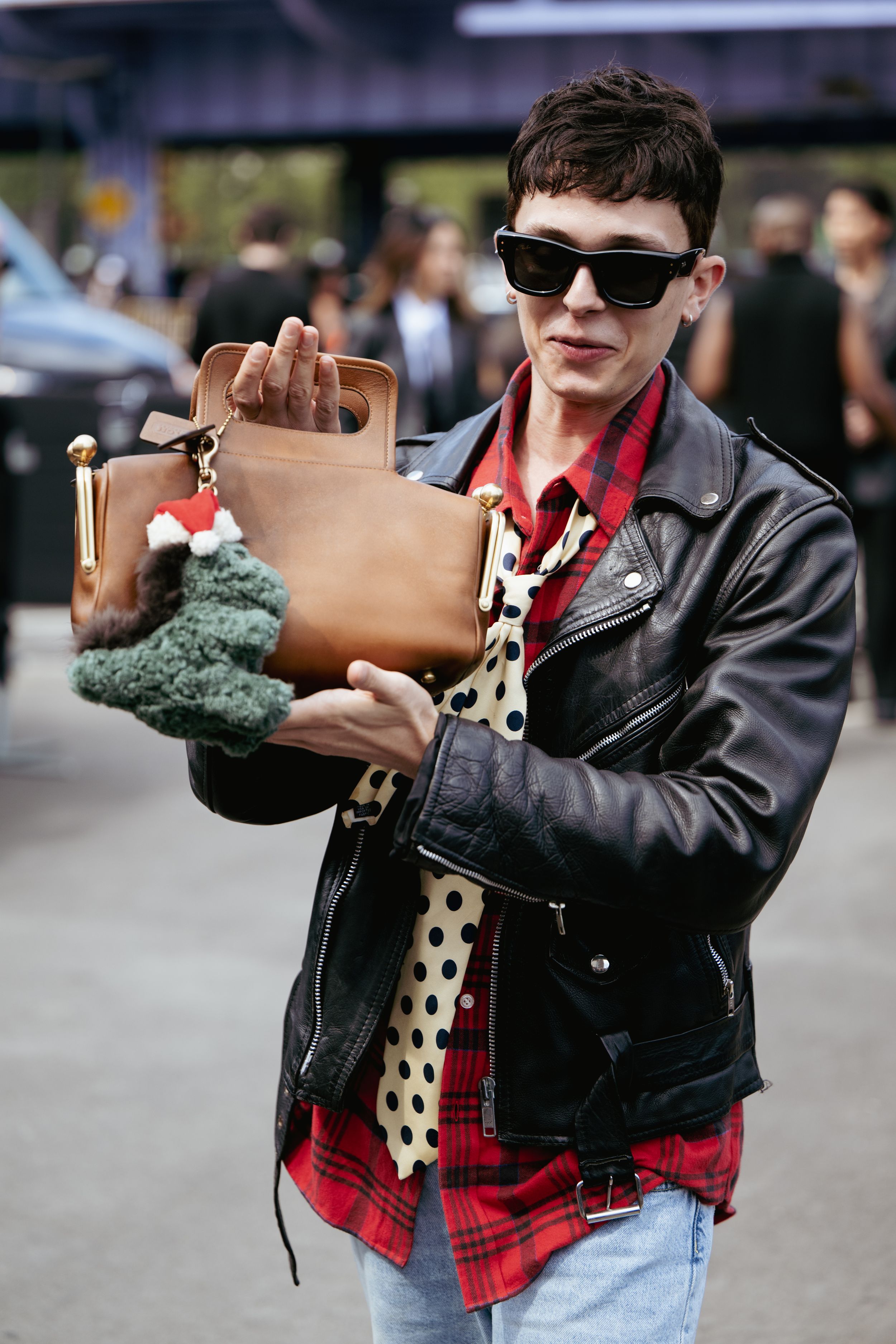 Street style fashion with leather jacket, plaid shirt, polka dot tie and brown leather bag on urban sidewalk.
