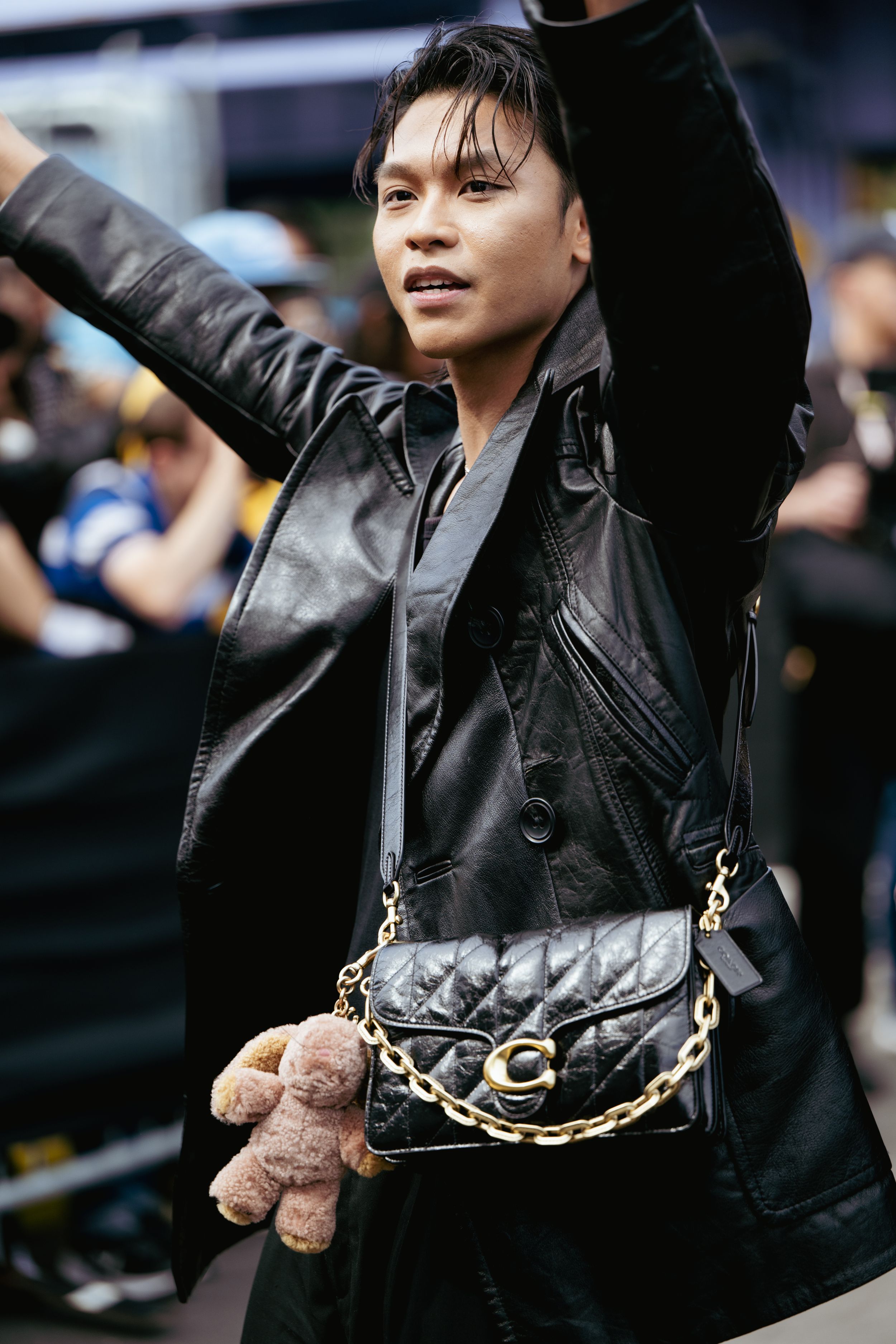 Person in black leather jacket with chain details and plush toy accessory poses against blurred background.