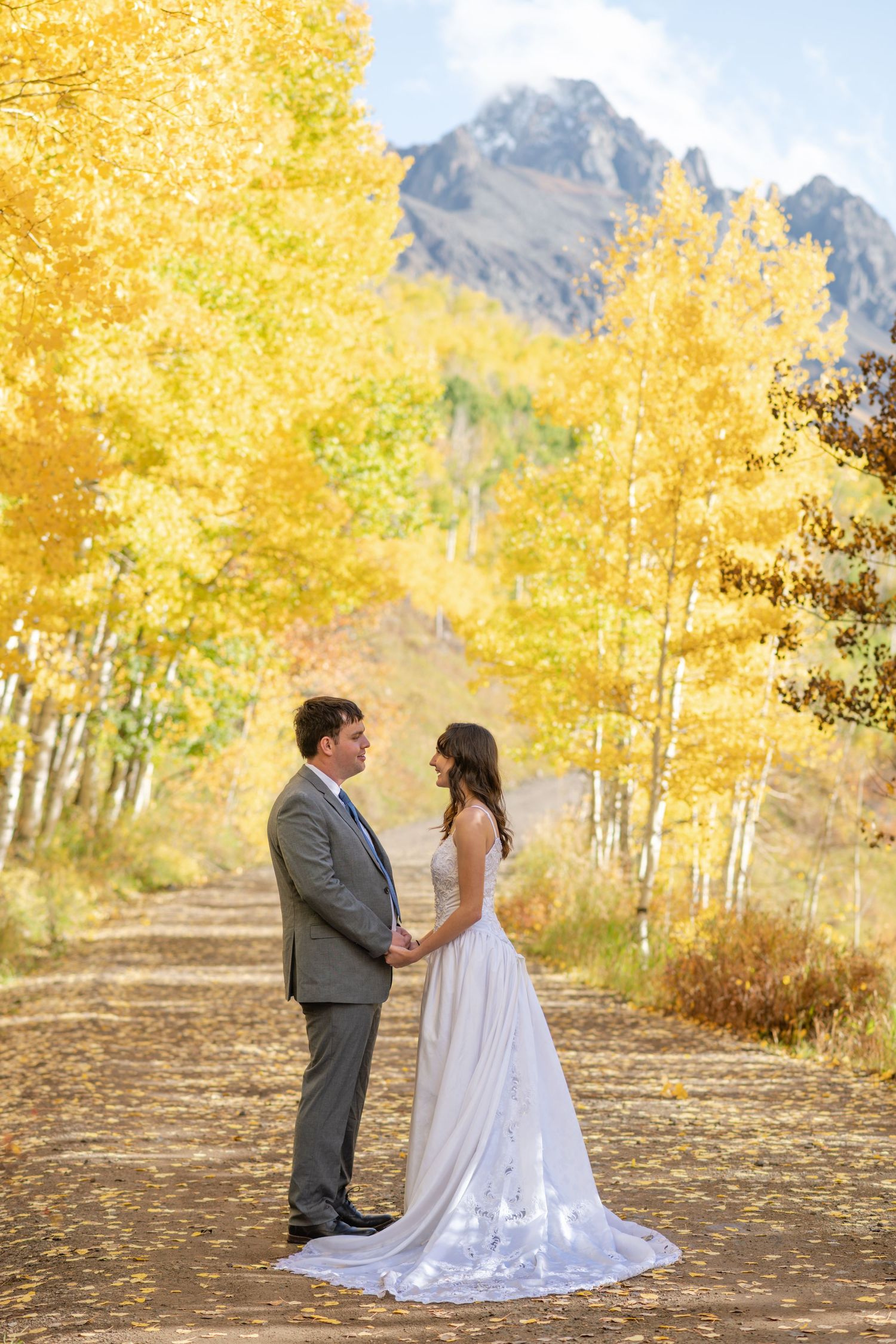 A bride and groom stand in the middle of a road lines with golden aspen in sunlight. They are holding hands facing each other.