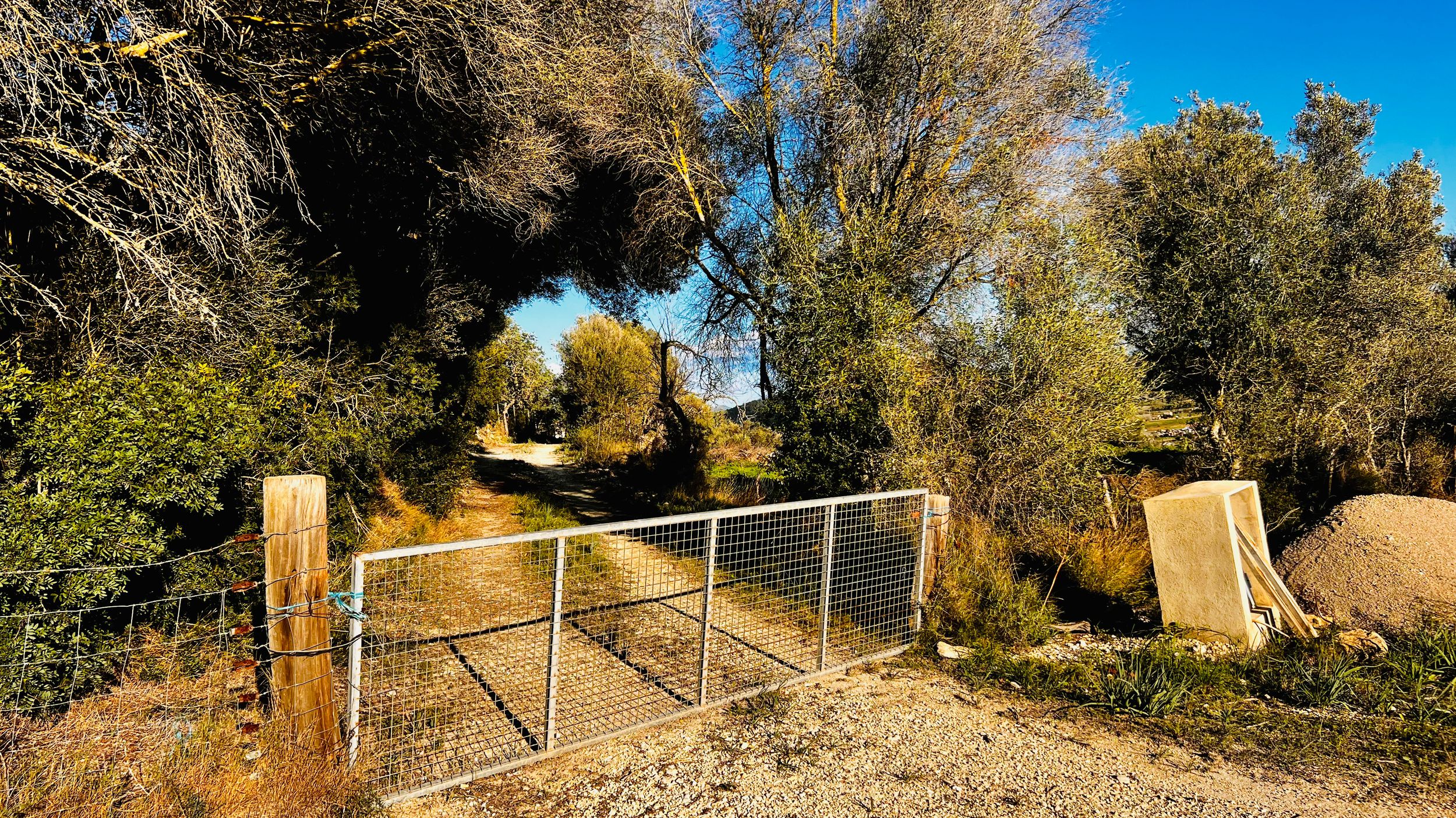 A winding dirt trail through autumn trees curves along a hillside with scattered wooden posts and fencing.