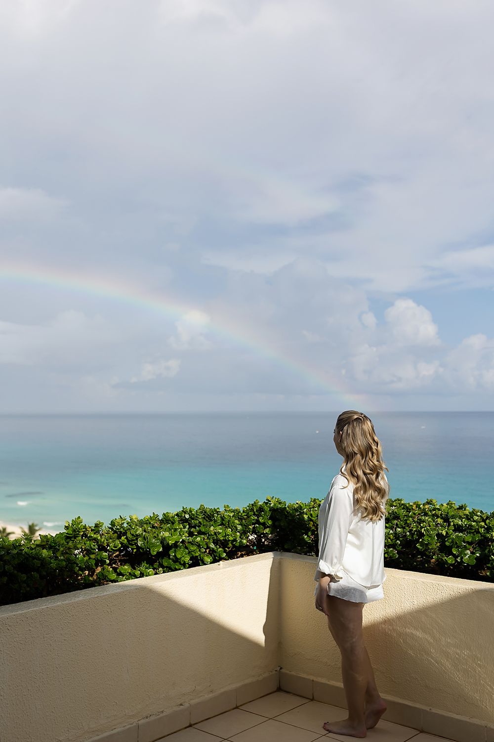 A rainbow appears over the ocean as someone looks out from a beige balcony with tropical plants.