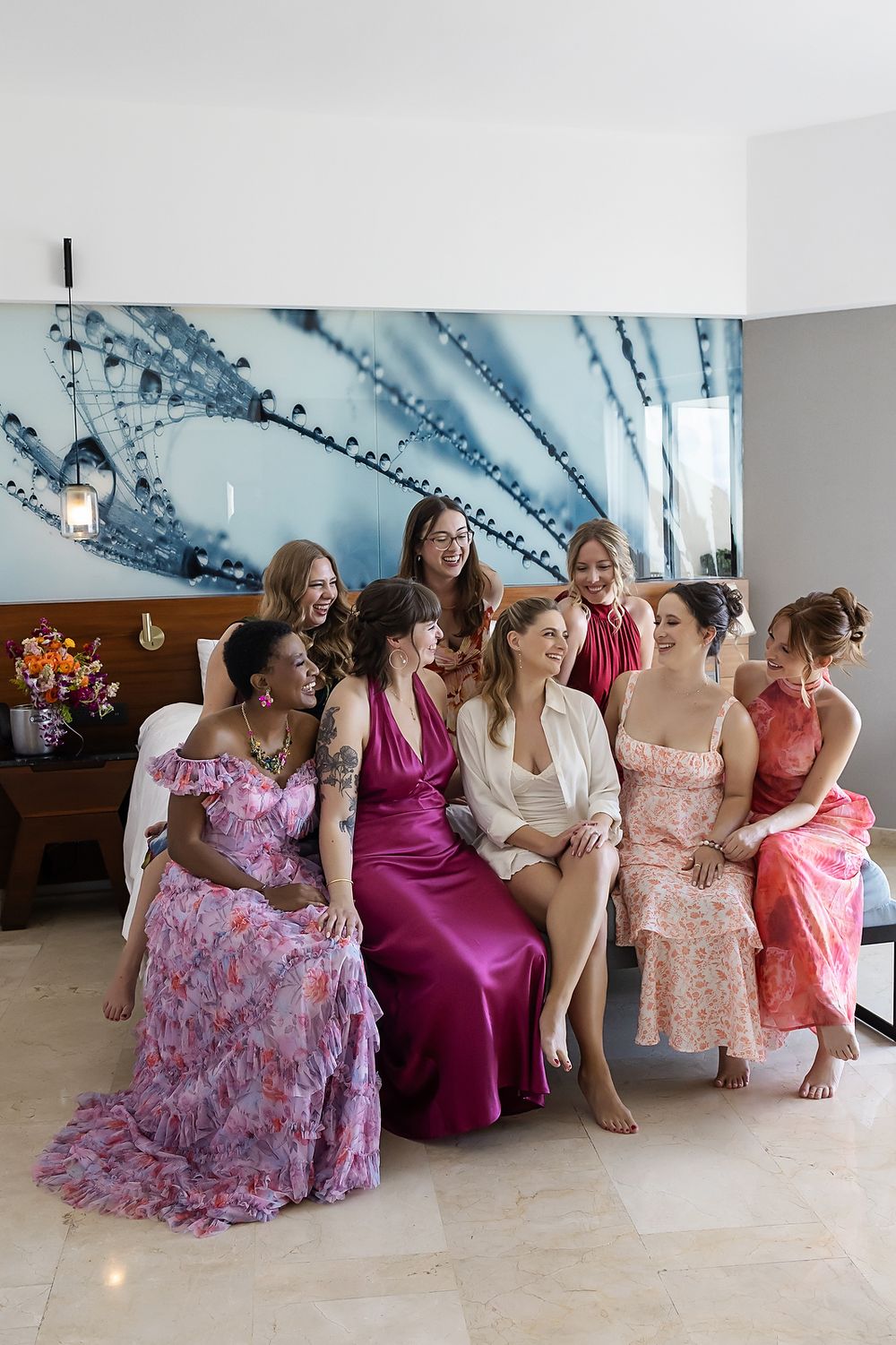 A group in formal dresses sits together on the floor in a bright room with floral and burgundy gowns.