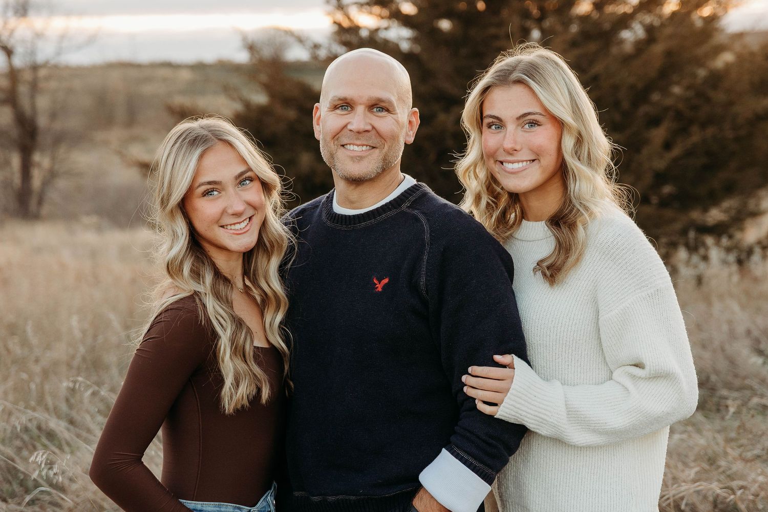A family wearing coordinated brown and cream outfits poses together in a sunlit outdoor setting during autumn.