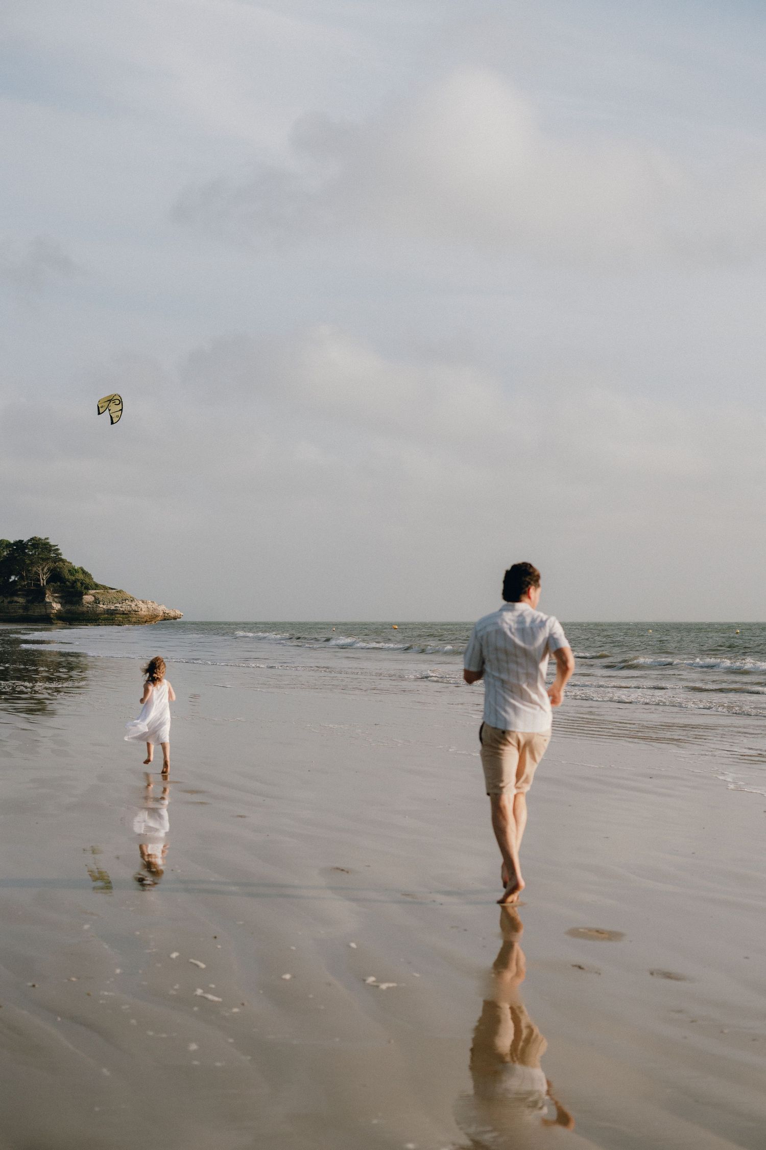 Papa court après sa fille sur la plage, cerf volant au fond.
