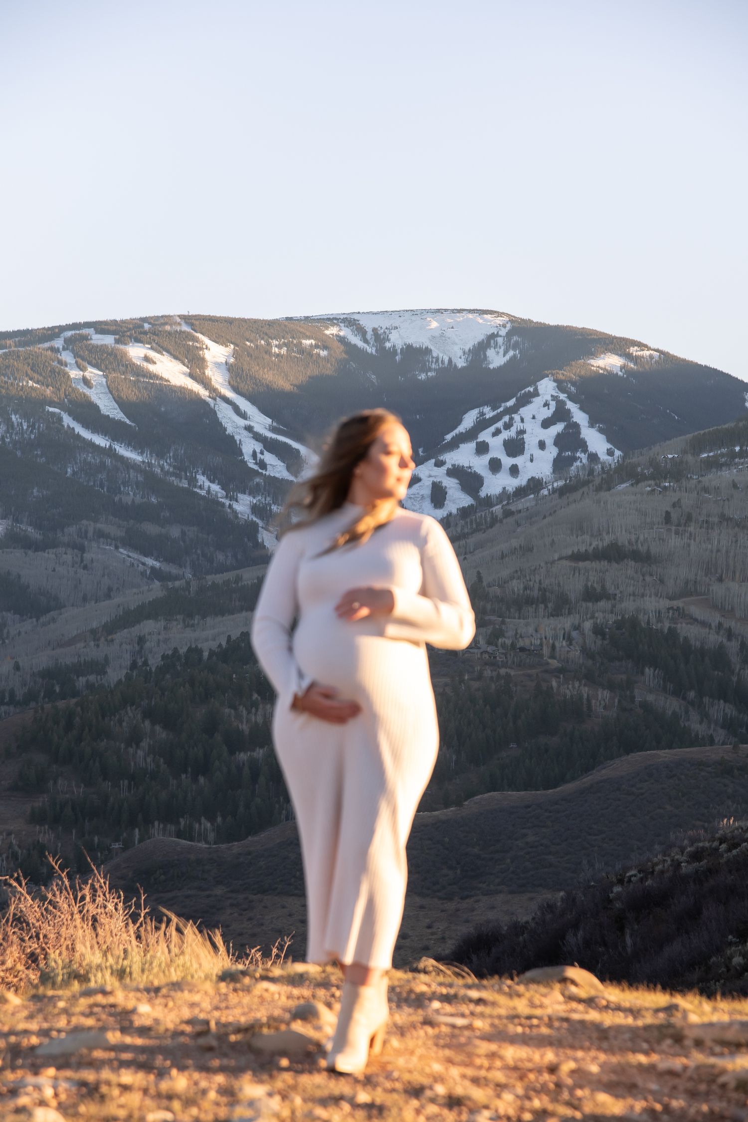 Artistic maternity photo session of a woman holding her pregnant belly while standing on top of a mountain during golden hour in Vail.
