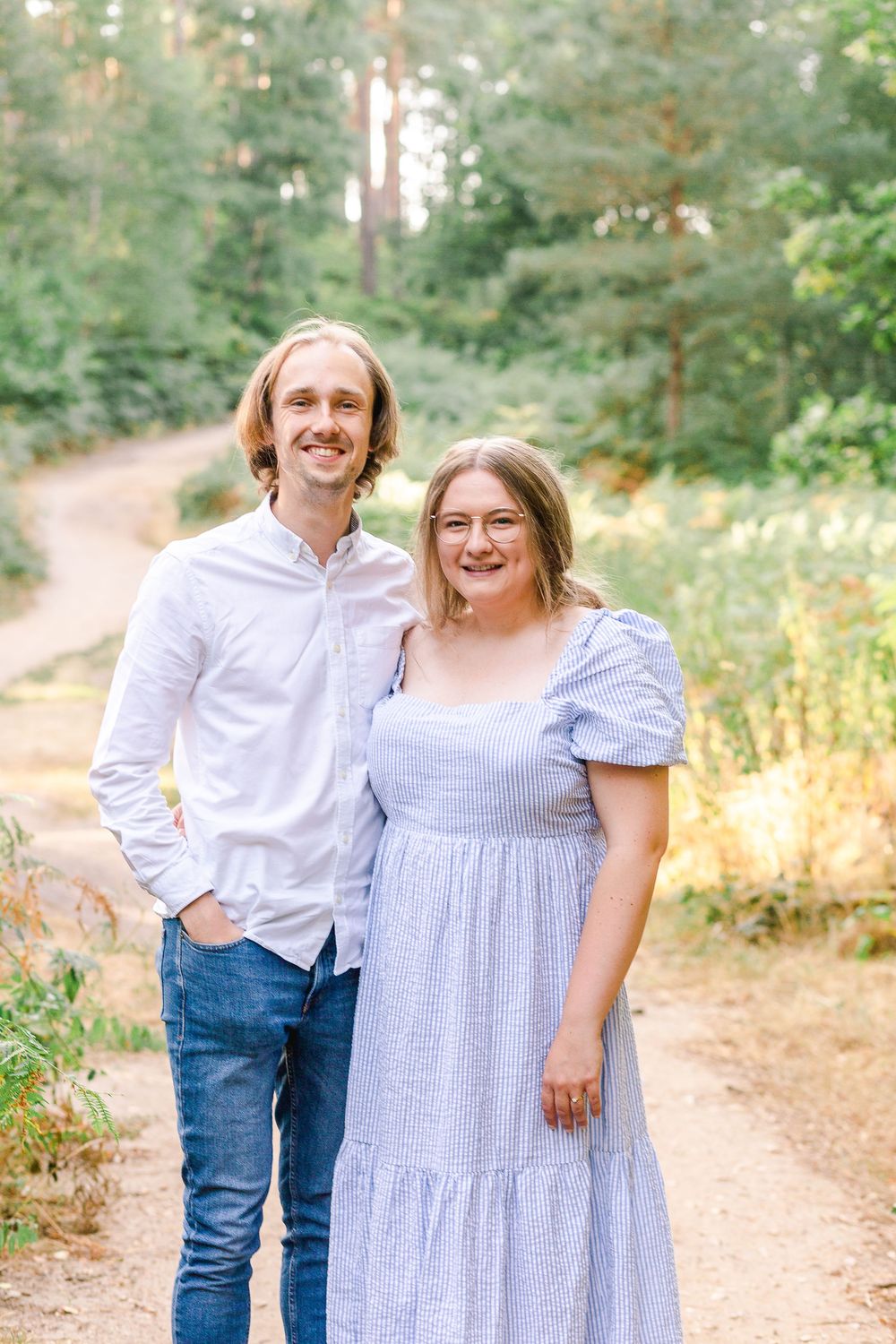 A couple wearing coordinating blue and white outfits poses together on a sunlit forest path during an outdoor photoshoot.