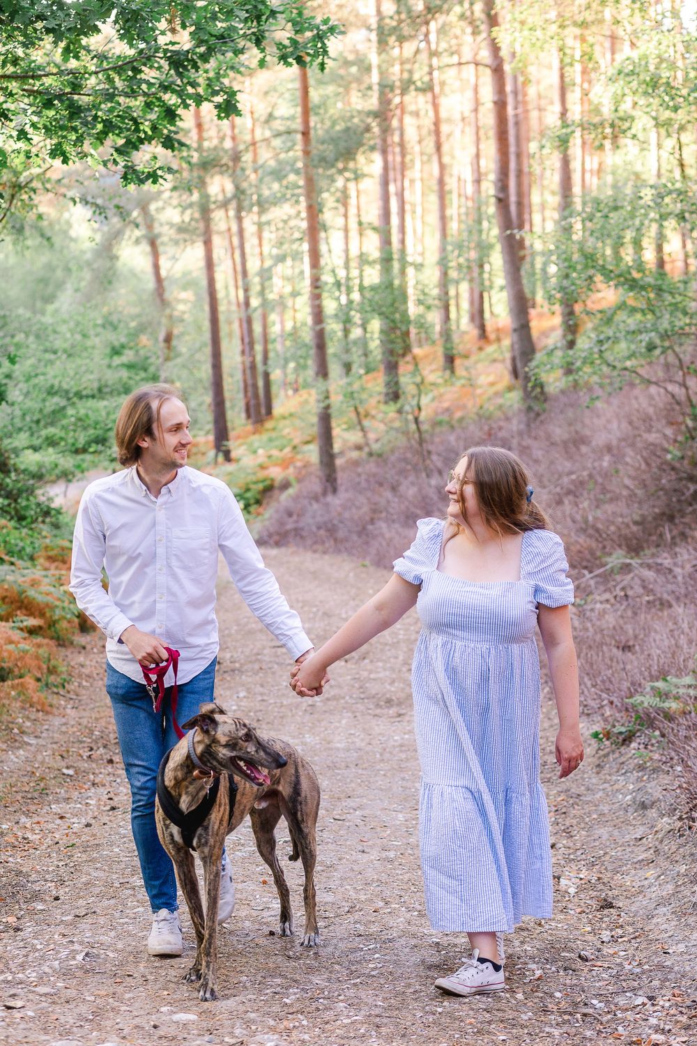 A couple in white outfits walks their brindle dog along a wooded trail during sunset.
