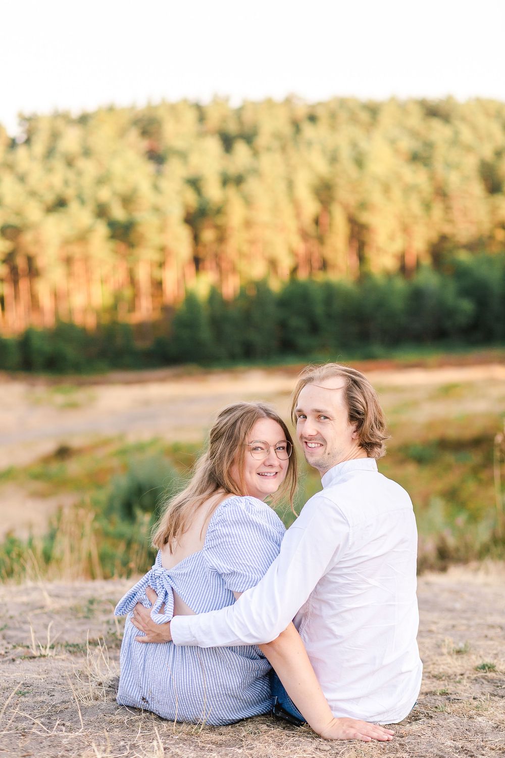 A couple embraces while sitting together on a hill overlooking a scenic forest at sunset.