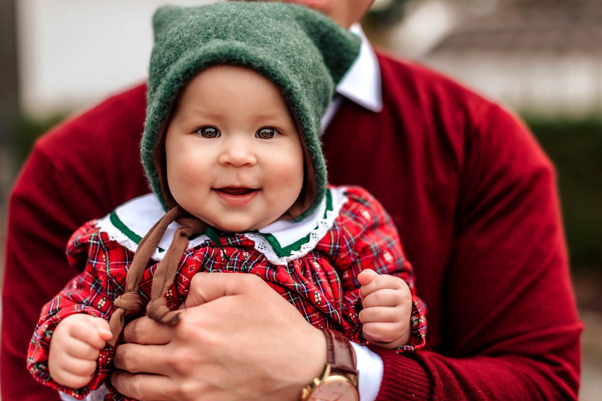 Nærbilde av en smilende baby på 7 måneder med grønn lue og rød kjole. Familie på julekortfotografering på Ålgård.