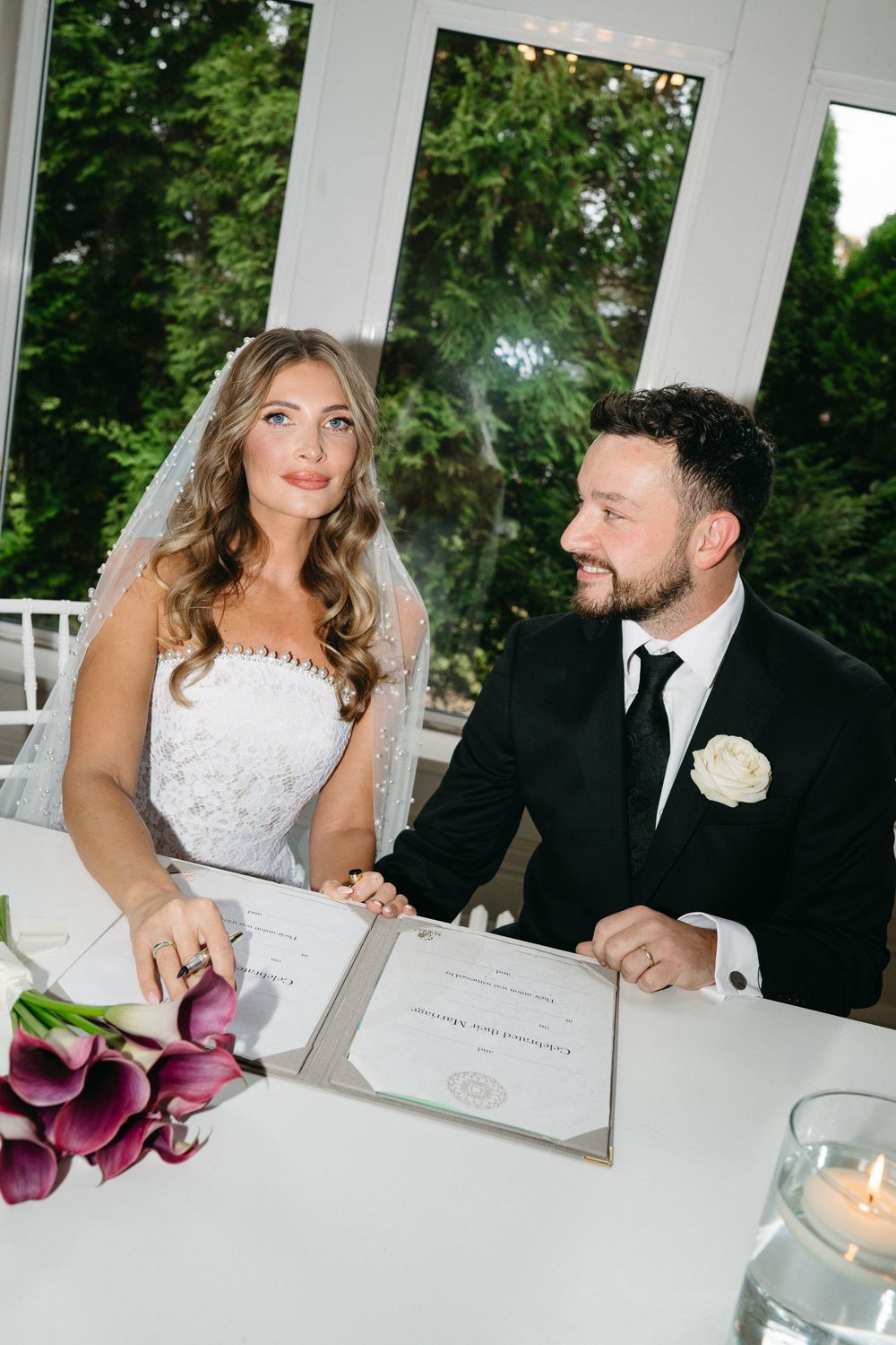 Newlyweds sign marriage documents at a white table with purple calla lily bouquet and candlelight.