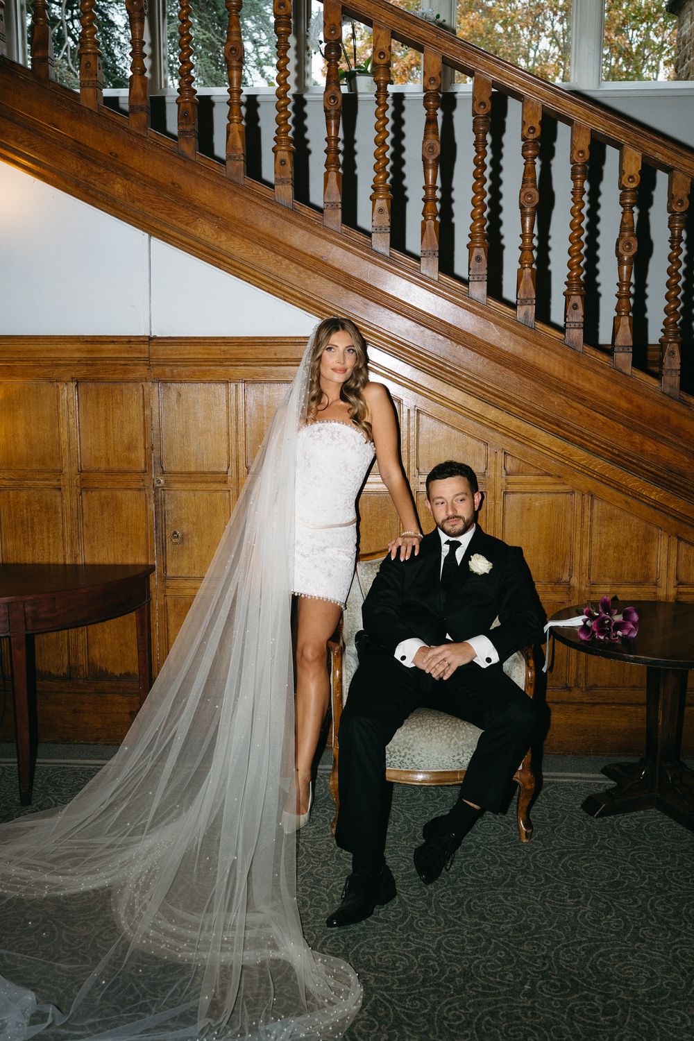 Wedding couple poses on elegant wooden staircase with long flowing veil trailing down the steps.
