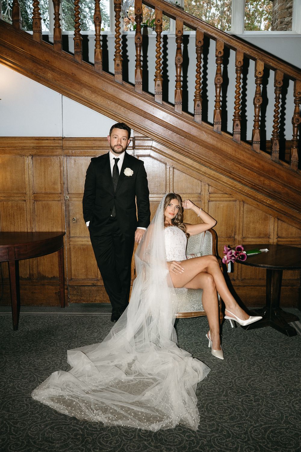 A bride in a white dress poses playfully on stairs next to a groom in a black suit near an elegant wooden banister.