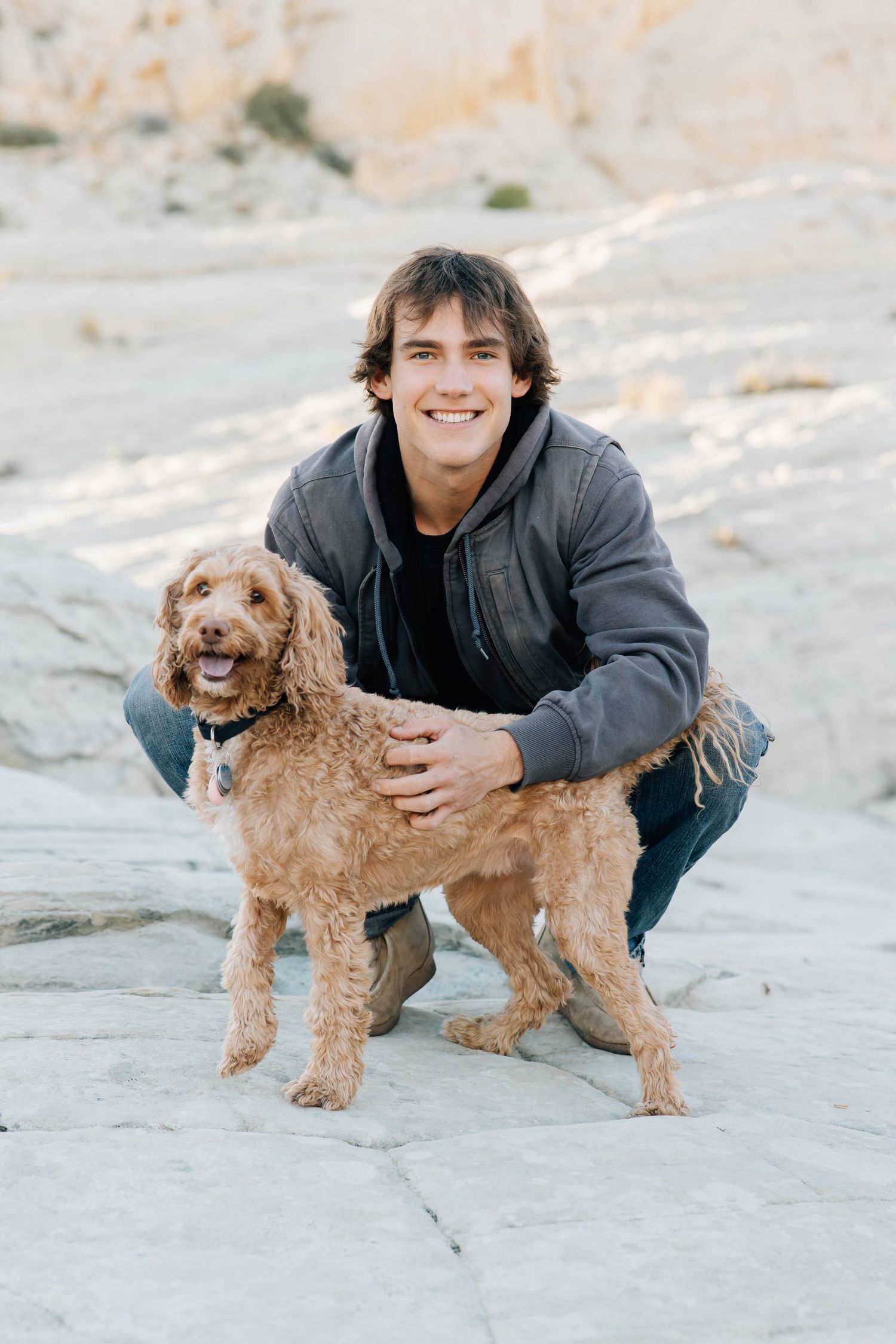 Smiling young man crouching with his fluffy golden doodle dog on rocky terrain outdoors.