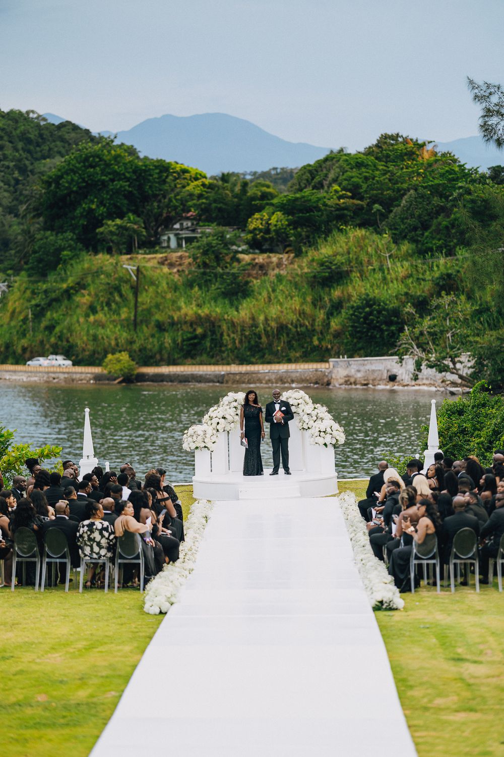 Panoramic view of an outdoor wedding ceremony setup with white decor against a lakeside backdrop and mountain scenery.