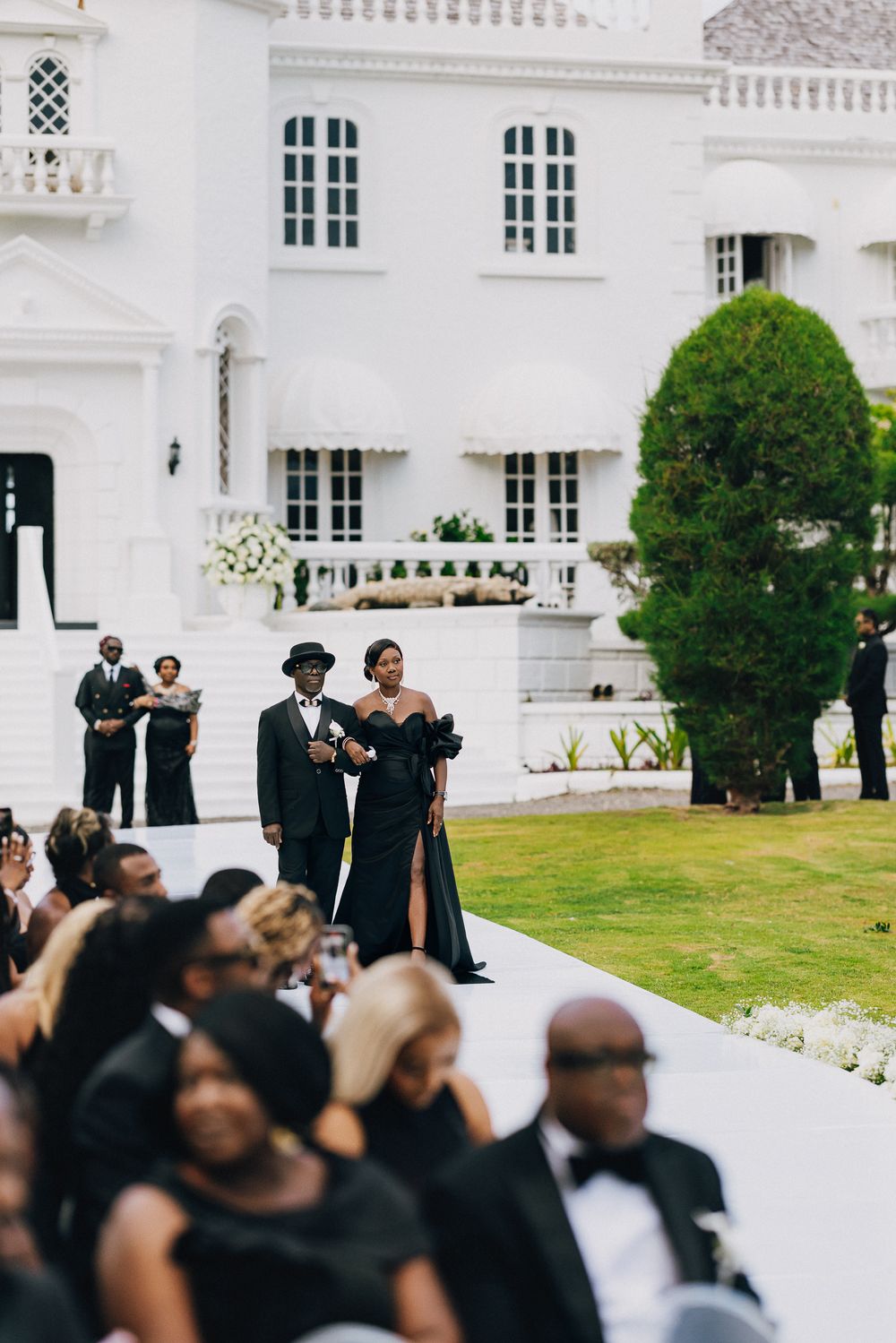 Wedding guests in formal black attire gather on a lawn outside an elegant white mansion with classic architecture.