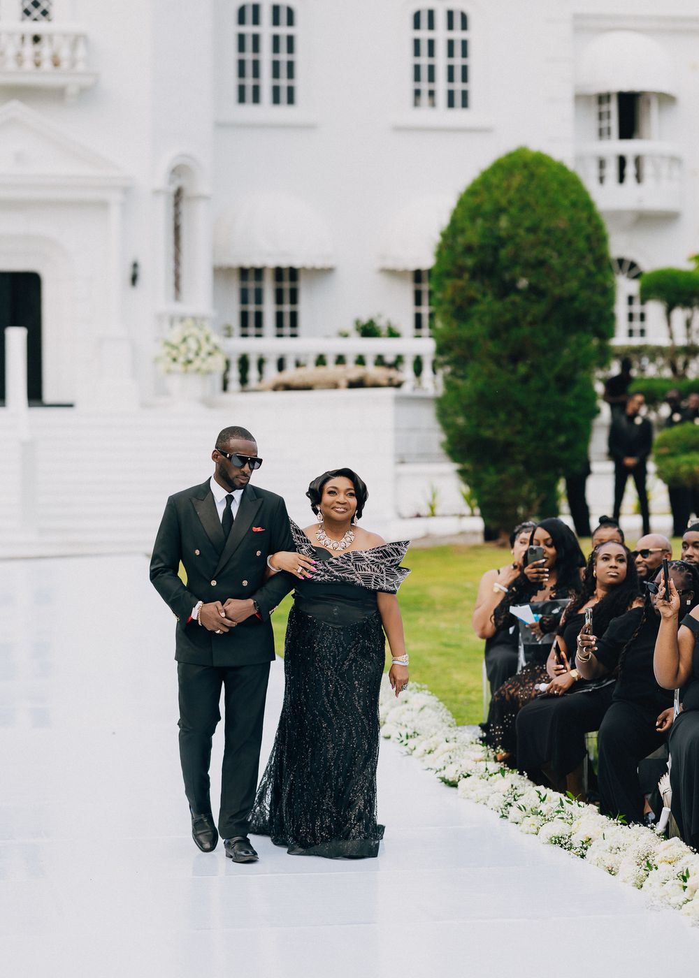 Wedding guests dressed in formal black attire walk down a white walkway next to a grand white building.