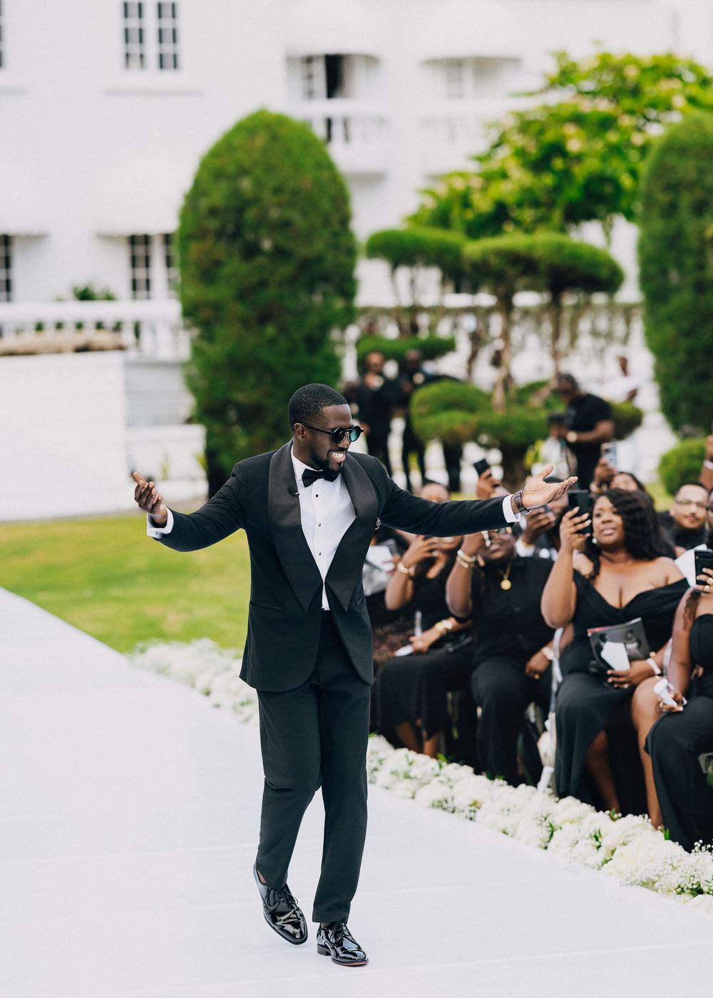 A person in a black tuxedo and bowtie dances joyfully on a white walkway while guests in black attire look on.