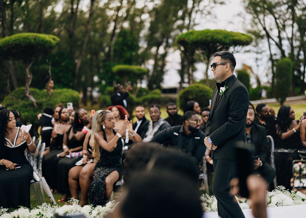 Outdoor wedding ceremony with guests in black attire gathered around ornate topiary trees and white floral arrangements.