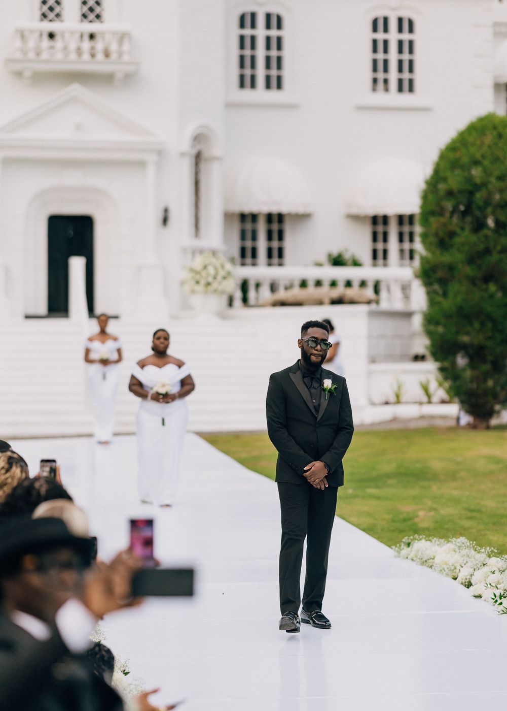 An outdoor wedding ceremony at an elegant white mansion with a white carpet aisle and lush green landscaping.