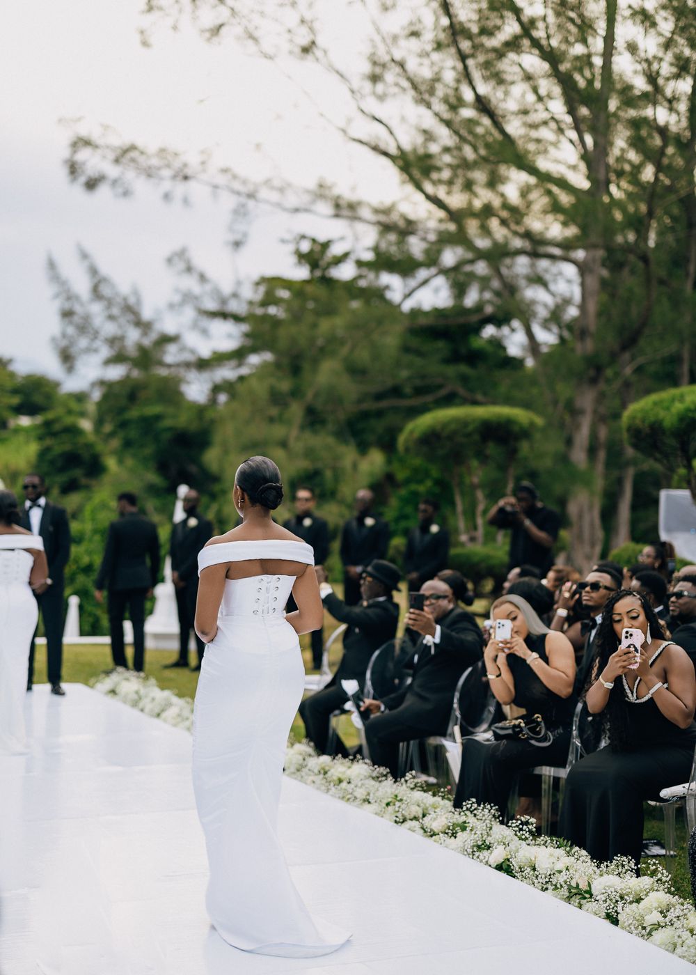 Wedding guests seated outdoors capture photos of the ceremony with their phones along a white floral-lined aisle.