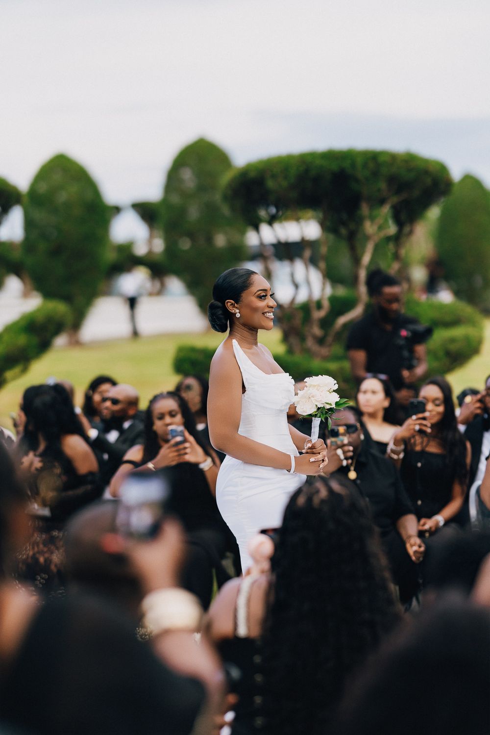 A bride in a white dress holds a bouquet while surrounded by wedding guests taking photos on their phones outdoors.